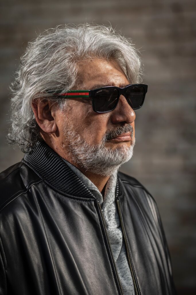 Man wearing sunglasses with a Gucci logo and a black leather bomber jacket, posing against a neutral backdrop, exuding a stylish and confident demeanor.