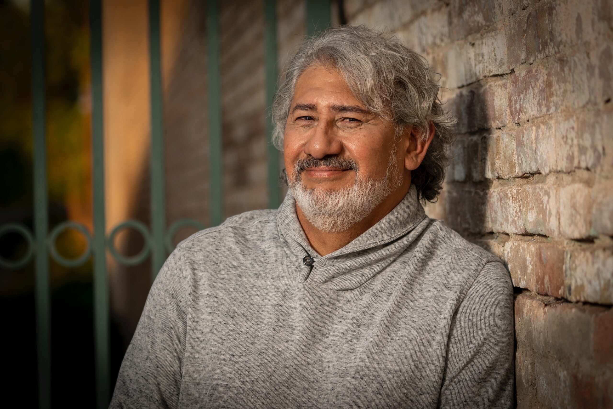 Smiling man with silver hair and a beard sits against a textured brick wall, wearing a gray sweater. The warm lighting creates a welcoming atmosphere.