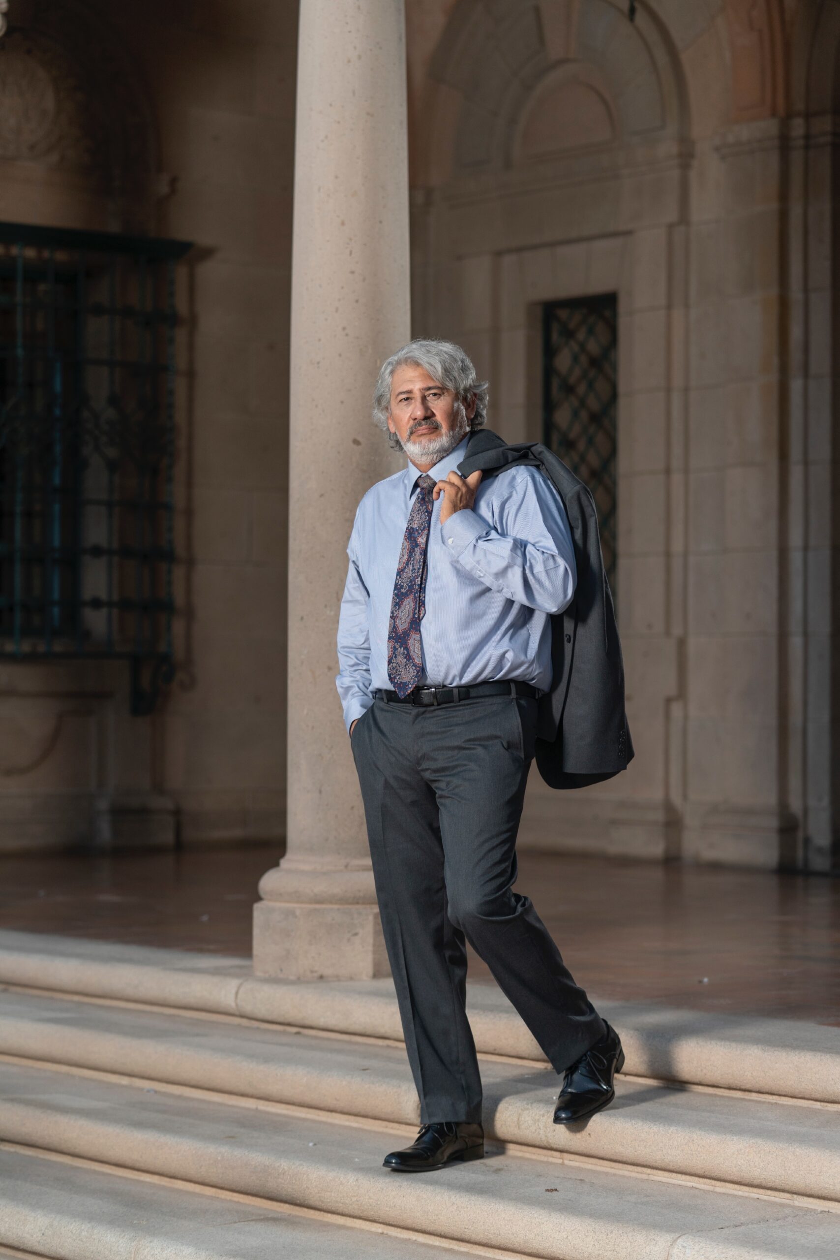A professional-looking man with gray hair stands confidently on steps, holding a suit jacket over his shoulder in a well-lit, elegant setting.